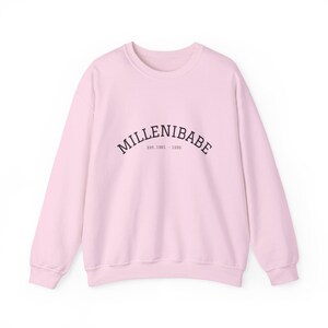 May include: A light pink long-sleeve crewneck sweatshirt featuring the black text MILLENIBABE arched across the chest, with the smaller text EST. 1961 - 1996 centered directly underneath the main word in a simple, clean font style.