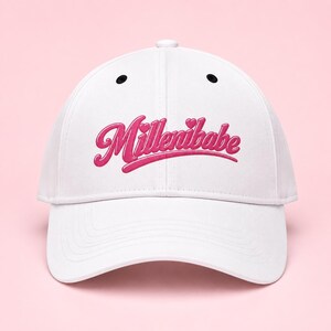 May include: A white baseball cap featuring the word Millenibabe embroidered in a bold, raised pink script font across the front panels. The design includes two small pink hearts above the letters and a curved pink line underlining the text.
