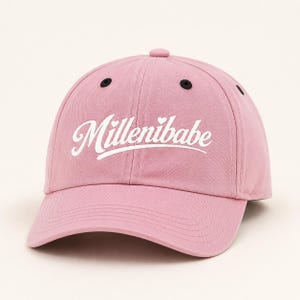 May include: A light pink baseball cap featuring the white embroidered text Millenibabe across the front. The cap has a curved brim, a structured crown with six panels, and small black eyelets at the top of each panel for ventilation.