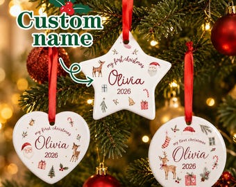 May include: Three white ceramic Christmas ornaments in star, heart, and circle shapes, each hanging from a red ribbon. The ornaments feature the text "my first christmas", the name "Olivia", the year "2026", and illustrations of a deer, Santa, and gifts.