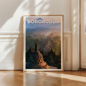 Może przedstawiać: Oprawiony wydruk artystyczny przedstawiający krajobraz świątyni Borobudur w Indonezji o wschodzie słońca. Obraz ukazuje kamienne stupy na pierwszym planie, bujne zielone drzewa oraz odległy szczyt góry pod miękkim, zamglonym niebem.