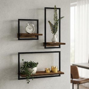 May include: A set of three wall-mounted shelves featuring black metal rectangular frames and dark brown wooden planks. The shelves hold a white analog clock, a green fern in a white vase, a potted green plant, and two small lit candles.
