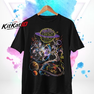 May include: A black short-sleeve t-shirt featuring a vibrant space-themed graphic print with two characters in a battle pose, planets, and the text "RINGS OF SATURN" in a stylized font. The shirt is displayed on a wooden hanger against a white background.