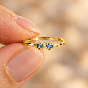 Puede incluir: Un anillo de oro con un diseño de símbolo de infinito. El bucle del infinito está acentuado con dos pequeñas piedras preciosas en forma de pera: una piedra de color azul verdoso a la izquierda y una piedra azul a la derecha, engastadas en una banda de oro pulido.
