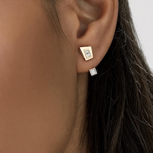 May include: A gold-toned ear jacket earring featuring a trapezoidal stud with two small rectangular clear stones and a separate smaller square clear stone stud positioned below the earlobe, creating a layered and geometric appearance.