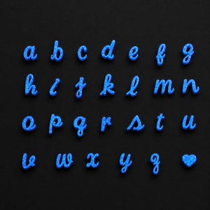 May include: A set of lowercase alphabet letters from a to z and a heart shape, all crafted in a bright blue material with a speckled, iridescent finish. Each letter is rendered in a cursive script font and arranged in four horizontal rows.