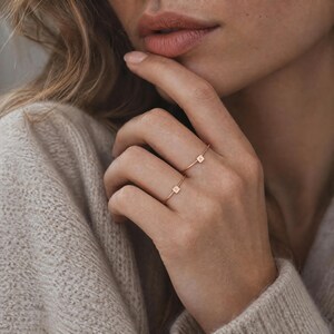以下が含まれることがあります： 小さな正方形のチャームが付いた2本の細いローズゴールドのリング。1つのチャームには文字Aが、もう1つには文字Kが刻印されています。リングは繊細なバンドデザインで、2本の指にそれぞれ装着されています。