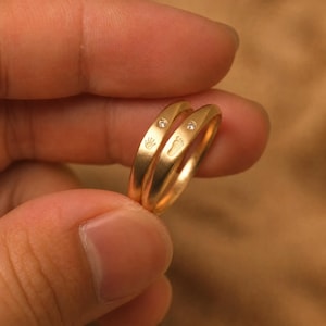 May include: Two gold rings with a smooth, polished finish. One ring features a small engraved handprint and a single embedded clear gemstone. The second ring displays a small engraved footprint and a single embedded clear gemstone.