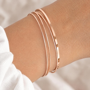 Könnte beinhalten: Ein Set aus drei dünnen Armreifen aus roségoldfarbenem Metall. Die Armreifen haben eine glatte, polierte Oberfläche und ein schlichtes, kreisförmiges Design. Jeder Armreif hat eine gleichmäßige, schmale Breite für einen Stapel-Look.