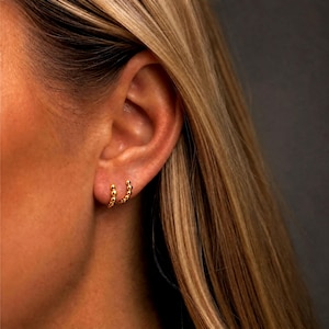 May include: A small gold hoop earring featuring a series of tiny, uniform spherical beads along the outer edge. The hoop is secured through the earlobe, showcasing a polished metallic finish that reflects light across its rounded, beaded surface.