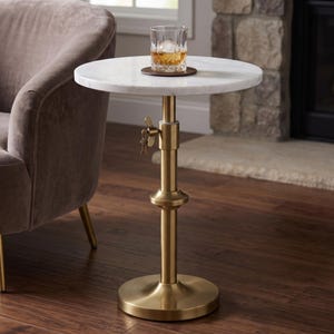 May include: A round side table with a white marble top and a brass-finished metal pedestal base. The central column features an adjustable height mechanism with a wing nut handle. A glass of amber liquid sits on a dark brown circular coaster.
