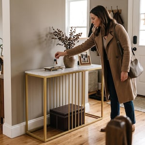 May include: A rectangular console table featuring a white marble top and a gold-toned metal frame with vertical bars. The table includes a small rectangular tray on top, a brown decorative vase with dried branches, and a dark brown storage box.