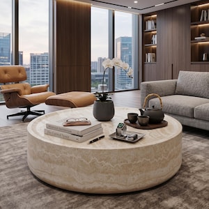 May include: A large, round, beige travertine coffee table with a natural veined texture. On the surface are two stacked books titled Minimalist Architecture and The Curated Space, a pair of glasses, a black pen, a potted white orchid, a smartwatch, a car key, and a dark teapot with two matching cups on a wooden tray.