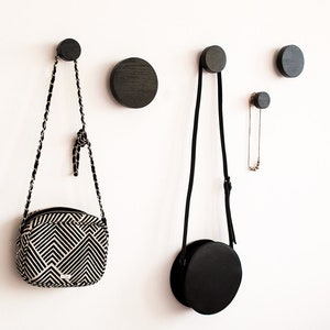 Op de afbeelding: Vijf zwarte houten haken gemonteerd aan een witte muur. Twee tassen hangen aan de haken, een met een zwart-wit geometrisch patroon en de andere een effen zwarte cirkel. Een gouden kettingketting hangt aan een derde haak.