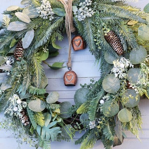 May include: A decorative wreath featuring a variety of green foliage, such as pine and eucalyptus, with brown pine cones and small white flowers. Two copper bells with heart shapes hang from a twine loop. A seasonal, natural design.