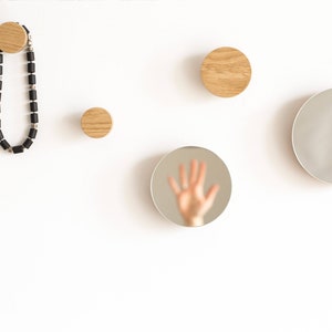 May include: Four wooden wall hooks with a round shape and a natural wood finish. A black beaded necklace hangs from one of the hooks. A round mirror reflects a hand.