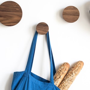 Peut inclure: Trois crochets muraux en bois avec un design rond de couleur noyer. Un sac fourre-tout bleu est suspendu à l'un des crochets, et un débardeur gris est suspendu à l'autre. Un casque de vélo bleu est sur le mur à droite.