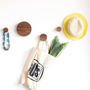 Könnte beinhalten: Runde Wandhaken aus Holz mit natürlicher Holzoptik. An den Haken hängen eine Halskette, eine Tragetasche, ein Bund Lauch und ein Strohhut.