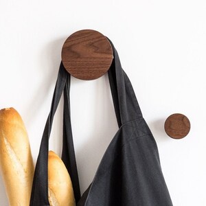 Può includere: Una borsa tote nera con una grafica bianca appesa a un gancio da parete in legno. Due baguette sono all'interno della borsa.