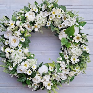 May include: A circular floral wreath composed of white roses, daisies, and hydrangeas, with green leaves. The wreath is displayed against a light grey wooden backdrop, offering a fresh and natural appearance.
