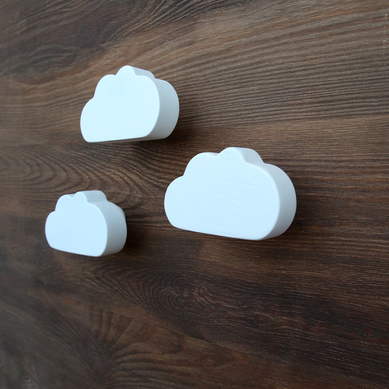 May include: Three white wooden cloud-shaped wall hooks. The hooks are mounted on a dark brown wooden surface.