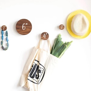 Op de afbeelding: Vier houten wandhaken met witte nummers erop, een haak van 5 cm, een haak van 10 cm, een haak van 15 cm en een haak van 5 cm. Een witte boodschappentas met zwarte tekst die "Gus" zegt, hangt aan de haak van 10 cm. Een ketting van blauwe en groene kralen hangt aan de haak van 5 cm. Een gele strohoed hangt aan de muur.