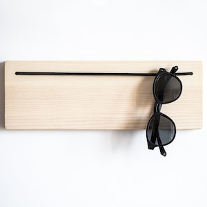 Peut inclure: Un porte-lunettes en bois à fixer au mur avec un cordon élastique noir. Une paire de lunettes de soleil noires est suspendue au cordon.