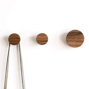 May include: Three round wooden wall hooks in different sizes. The largest hook is on the right, the smallest is on the left, and the middle-sized hook is in the centre. A brown strap is hanging from the leftmost hook.