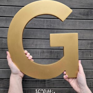 Puede incluir: Una letra "G" dorada de aproximadamente 41 cm de altura. La letra se sostiene sobre un fondo de madera oscura. El texto "16" letter" está escrito en cursiva en la parte inferior de la imagen.