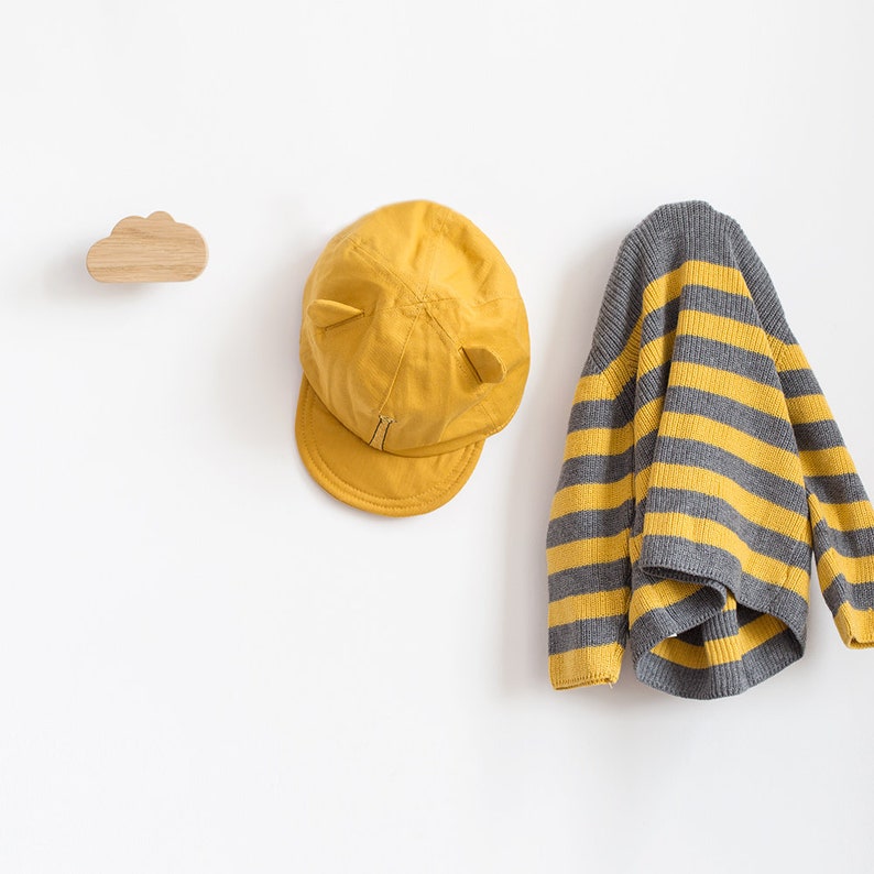 May include: A yellow baseball cap with ear flaps and a grey and yellow striped sweater hanging on a wooden cloud-shaped hook against a white background.