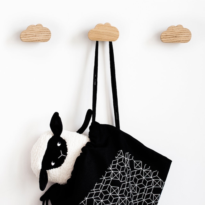 May include: Three wooden cloud-shaped hooks mounted on a white wall. A black tote bag with a white geometric pattern hangs from one of the hooks. A black and white stuffed sheep is partially inside the bag.