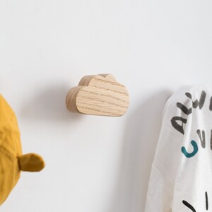 May include: A wooden cloud-shaped wall hook in a light wood tone. The hook is mounted on a white wall and has a garment hanging from it.