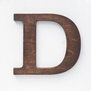 May include: A dark brown wooden letter D, standing upright on a white background.