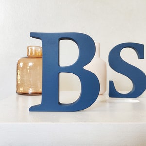 May include: Two wooden letters, "B" and "S", painted in a dark blue colour. The letters are standing on a white surface.