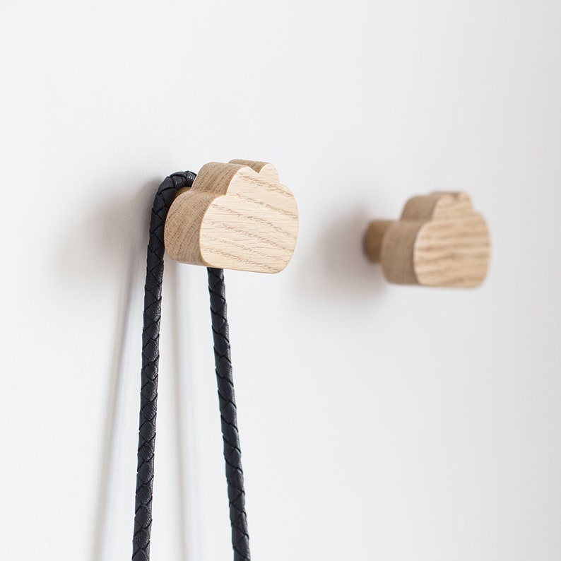 May include: Two wooden cloud-shaped wall hooks, one with a black braided cord hanging from it. The hooks are mounted on a white wall.
