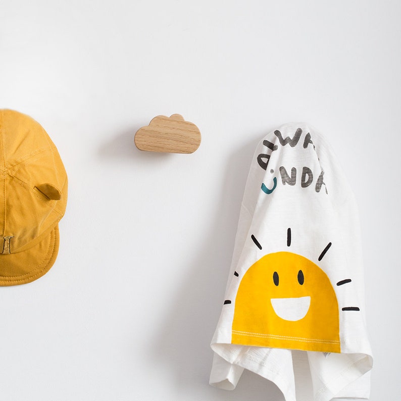 May include: A wooden cloud-shaped hook mounted on a white wall with a yellow baseball cap and a white towel with a yellow sun and the text "Always Wonder" hanging from it.