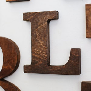 May include: A close-up of a wooden letter L, painted in a dark brown color. The letter is made of plywood and has a rustic, handcrafted look.