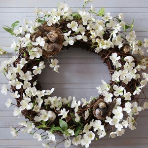 May include: A spring wreath made of twigs and white blossoms with small bird nests and eggs.