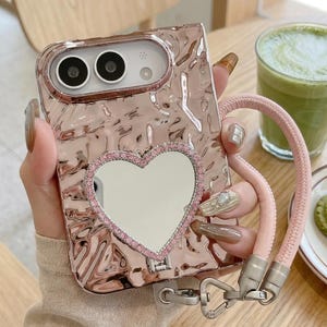 May include: A rose gold phone case with a metallic, hammered texture. The case features a heart-shaped mirror framed by small pink rhinestones on the back. A detachable, light pink braided wrist strap is attached to the bottom with a silver clasp.
