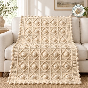 May include: A cream-colored crochet blanket featuring a repeating square pattern with a central raised bobble stitch in each square. The blanket is bordered by a textured edge with small pom-poms and includes a circular logo in the corner.