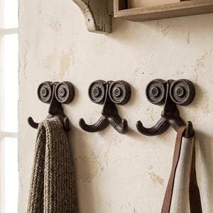 May include: Three dark brown cast iron wall hooks, each shaped like a face with two circular eyes and a mustache-like double hook. The hooks are mounted horizontally, with a textured knit fabric hanging from the left hook and a leather strap on the right.