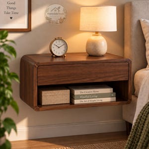 May include: A wall-mounted floating nightstand crafted from dark brown wood with a single drawer and an open storage shelf. The shelf contains a rectangular box and three stacked books titled The Creative Home, Mindful Living, and The Art of Simple Living.