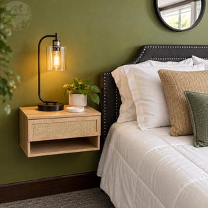 May include: A light wood floating nightstand with a cane drawer front and an open lower shelf. A black metal lamp with a clear glass cylindrical shade sits on top next to a small green potted plant in a white ribbed ceramic pot.