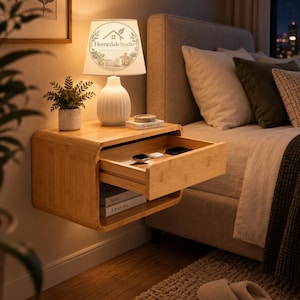 May include: A wall-mounted light wood floating nightstand with a single pull-out drawer containing a smartwatch and small electronic accessories. Below the drawer is an open storage shelf holding two books. A white table lamp sits on top.