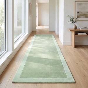 Puede incluir: Una alfombra de pasillo larga y rectangular en un color verde salvia claro sólido. La alfombra presenta un borde distintivo de un verde ligeramente más oscuro que enmarca la sección central rectangular, creando un diseño limpio en el suelo.