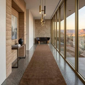 May include: A long hallway featuring a brown runner rug on a speckled gray floor, a slim console table with a black vase, a dark curved bench against a stone wall, and a cluster of cylindrical pendant lights hanging from the ceiling.