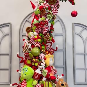 Könnte beinhalten: Ein hoher, verspielter Weihnachtsbaum, geschmückt mit roten und grünen Ornamenten, bonbonartigen Akzenten und Plüschfiguren, darunter ein grünes Wesen, eine blonde Figur und ein brauner Hund, auf einem runden, flauschigen grünen Sockel.
