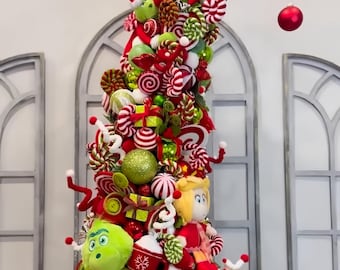 Pode incluir: Uma árvore de Natal alta e extravagante decorada com ornamentos vermelhos e verdes, detalhes temáticos de doces e personagens de pelúcia, incluindo uma criatura verde, uma figura loira e um cão castanho, sobre uma base circular de pelúcia verde.