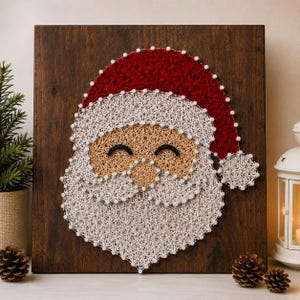 Pode incluir: Uma peça de arte com fios sobre uma placa de madeira castanho-escura representando o Pai Natal. O design utiliza fios brancos, vermelhos e beges enrolados em pregos prateados para formar o gorro, a barba, o bigode e o rosto.