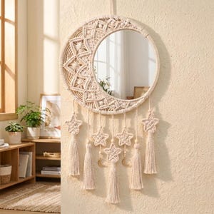 May include: A circular wall mirror framed by a cream-colored macrame crescent moon design. The piece features intricate knotted patterns, five hanging star-shaped macrame ornaments, and long tassels, creating a textured, layered wall hanging.